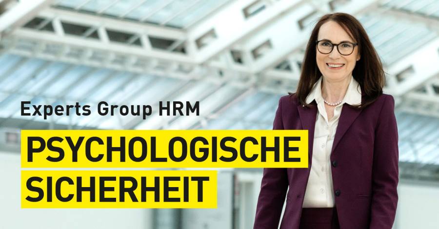 Person im Business-Outfit vor modernem Gebäude mit gelbem Schriftbanner „Psychologische Sicherheit“.