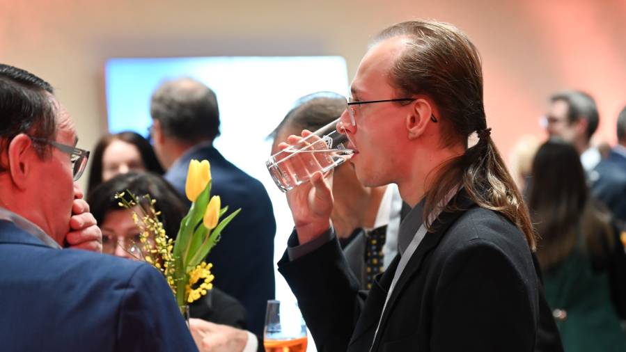 Mann mit Zopf trinkt Wasser, Menschen plaudern im Hintergrund miteinander